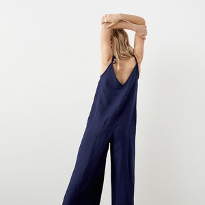 May include: A navy blue jumpsuit with wide legs and spaghetti straps. The jumpsuit has a V-neckline and tie details at the shoulders. The model is wearing white sneakers. The fabric appears to be linen or a similar lightweight material.