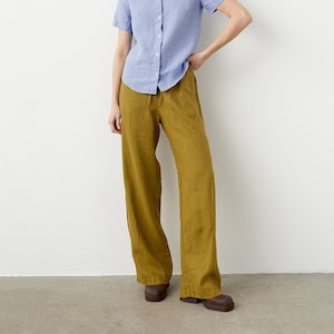 Puede incluir: Una persona viste una camisa abotonada azul claro de manga corta y pantalones de pierna ancha verde oliva. El atuendo se completa con zapatos marrones. La ropa parece ser de lino.