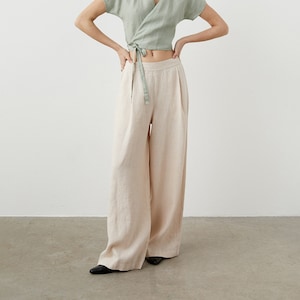 Puede incluir: Una mujer lleva una blusa de lino verde salvia con cuello en V y mangas cortas con un lazo a juego. Lleva pantalones anchos de lino beige.