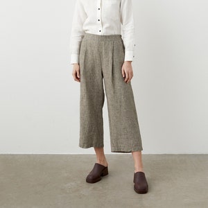 K&ouml;nnte beinhalten: Eine Frau tr&auml;gt ein wei&szlig;es Hemd mit schwarzen Kn&ouml;pfen und eine olivgr&uuml;ne, weit geschnittene, cropped Hose. Sie tr&auml;gt auch braune Lederclogs. Das Outfit ist schlicht und elegant.