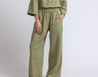 May include: A two-piece olive green linen outfit. The top has a mandarin collar, three-quarter sleeves, and a button closure. The pants are wide-legged with an elastic waistband. The model is wearing black sandals.