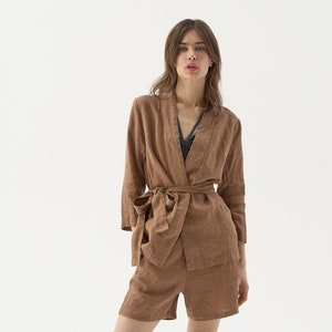 May include: A brown linen kimono-style jacket with a matching belt, paired with brown linen shorts. The jacket has wide sleeves and a wrap-around design. The shorts are high-waisted. The outfit is made of a lightweight, textured fabric.