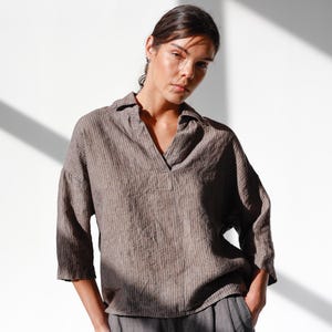 May include: A woman wearing a brown, striped linen shirt with a V-neck collar and three-quarter sleeves. She is also wearing gray linen pants. The shirt has a relaxed fit and the pants have a pleated front.