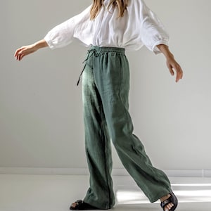 Puede incluir: Una mujer lleva una blusa de lino blanca de corte holgado y mangas largas. También lleva pantalones de lino verde oscuro holgados con cintura de cordón. Los pantalones tienen pierna ancha y están cortados en el tobillo. Lleva sandalias negras.