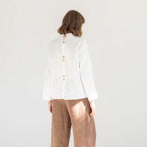 May include: A white linen blouse with long sleeves and wooden buttons down the back, worn with brown wide-leg trousers. The outfit is finished with black shoes. The ensemble suggests a relaxed, comfortable style, suitable for everyday wear.