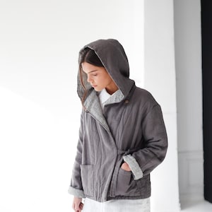 May include: A gray hooded jacket with a wrap-style closure and pockets. The jacket has a relaxed fit and is worn over a white shirt and dark blue pants. The sleeves are cuffed, revealing a lighter gray lining.