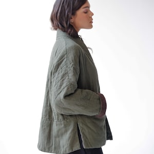May include: An olive green quilted jacket with brown cuffs. The jacket has a relaxed fit, a mandarin collar, and a side slit. The jacket is made of a soft, textured fabric.