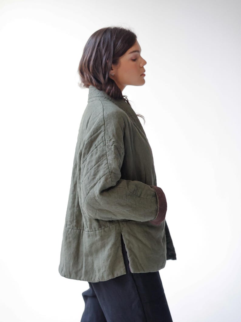 May include: An olive green quilted jacket with brown cuffs. The jacket has a relaxed fit, a mandarin collar, and a side slit. The jacket is made of a soft, textured fabric.