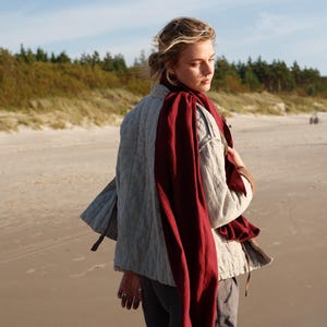 May include: A light gray quilted jacket with a burgundy scarf draped over the shoulders. The jacket has a relaxed fit and a subtle texture. The person is standing on a sandy beach with the ocean in the background.