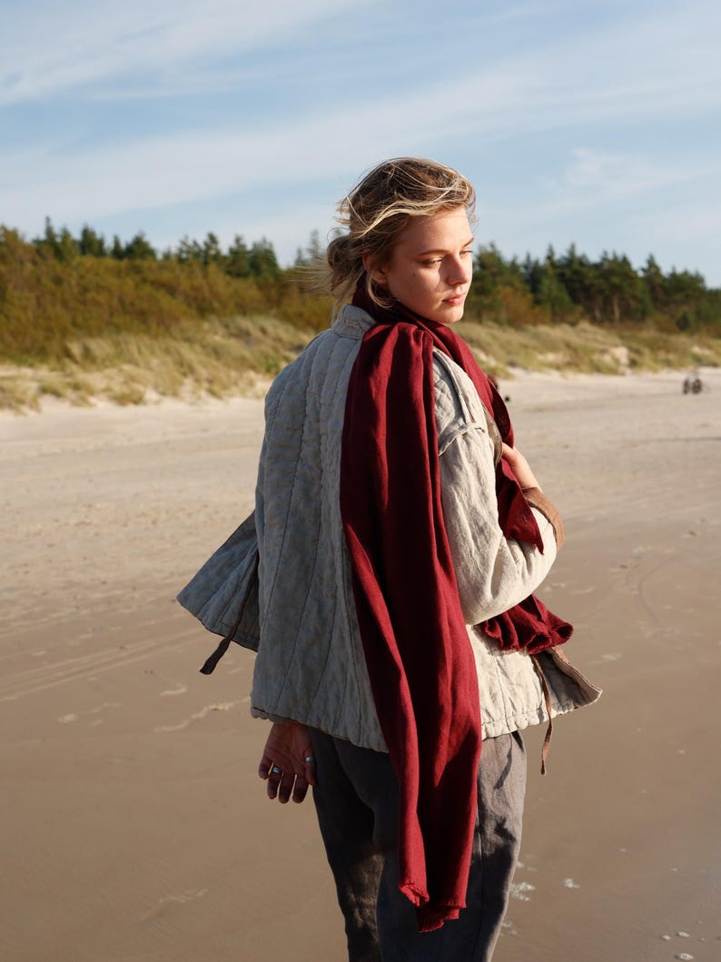 May include: A light gray quilted jacket with a burgundy scarf draped over the shoulders. The jacket has a relaxed fit and a subtle texture. The person is standing on a sandy beach with the ocean in the background.