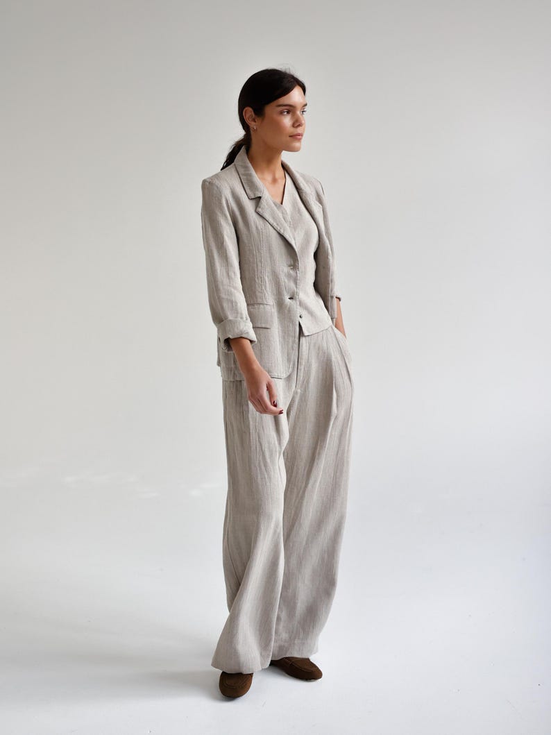 May include: A light beige linen suit, consisting of a blazer with three-quarter sleeves, a matching top, and wide-leg pants. The suit is worn with brown shoes. The outfit is made of a textured fabric and has a relaxed fit.