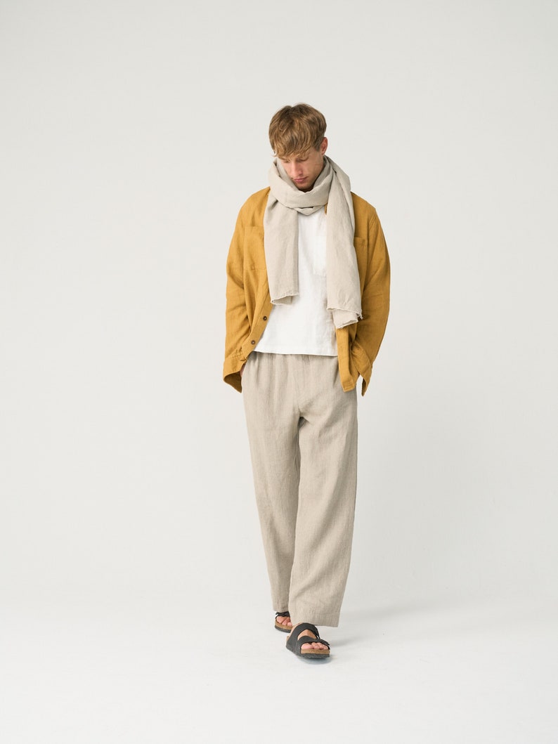 May include: A man wearing a mustard yellow linen jacket, a white t-shirt, beige linen pants, and a light gray linen scarf. He is wearing black sandals.