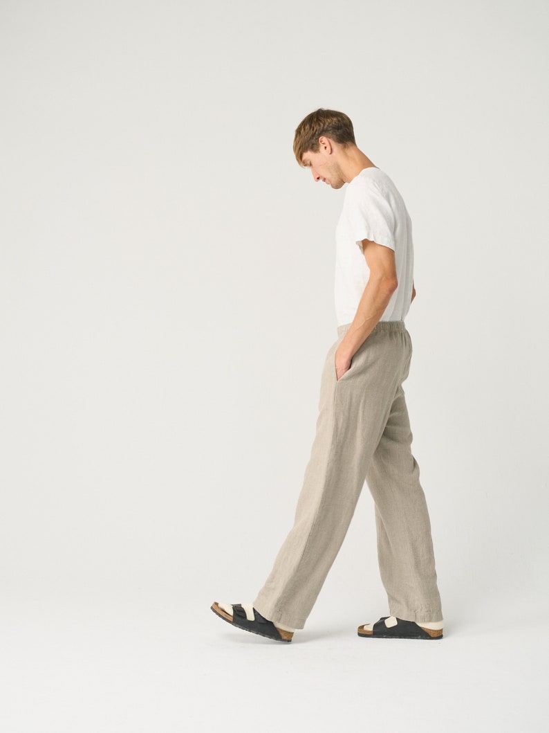 May include: A man wearing a white t-shirt and beige linen pants walks in a studio setting. He is wearing black sandals with white straps.
