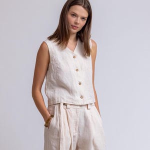 May include: A sleeveless, cream-colored linen top with a V-neck and wooden buttons. Paired with matching high-waisted, wide-leg linen pants. The outfit is a neutral, minimalist style, suitable for warm weather.