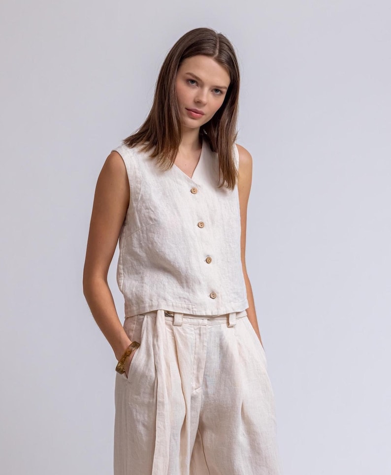 May include: A sleeveless, cream-colored linen top with a V-neck and wooden buttons. Paired with matching high-waisted, wide-leg linen pants. The outfit is a neutral, minimalist style, suitable for warm weather.