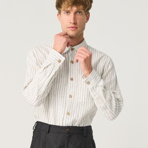 Op de afbeelding: Een cr&egrave;mekleurig shirt met lange mouwen, verticale zwarte strepen en houten knopen. Het shirt heeft een enkele borstzak en wordt gecombineerd met een zwarte broek. De outfit is eenvoudig en klassiek.