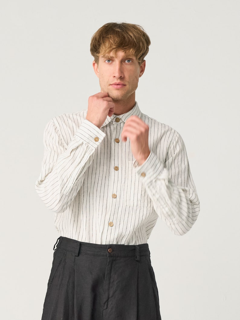 Op de afbeelding: Een cr&egrave;mekleurig shirt met lange mouwen, verticale zwarte strepen en houten knopen. Het shirt heeft een enkele borstzak en wordt gecombineerd met een zwarte broek. De outfit is eenvoudig en klassiek.