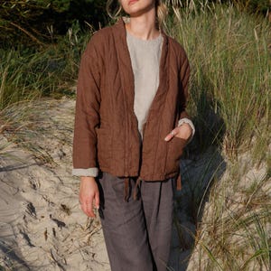 May include: A brown quilted jacket with a relaxed fit, paired with gray linen pants. The jacket features a V-neck design and pockets. The outfit is completed with a light-colored top underneath. The model is standing on a sandy beach.