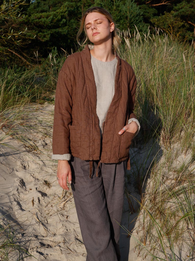 May include: A brown quilted jacket with a relaxed fit, paired with gray linen pants. The jacket features a V-neck design and pockets. The outfit is completed with a light-colored top underneath. The model is standing on a sandy beach.