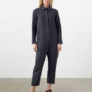 May include: A dark gray linen jumpsuit with long sleeves and a button-up front. The jumpsuit has a relaxed fit and a cropped leg.