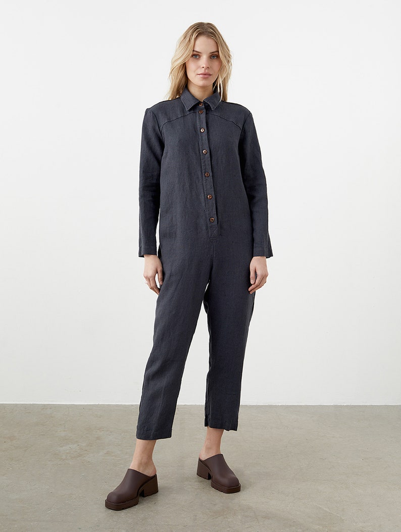 May include: A dark gray linen jumpsuit with long sleeves and a button-up front. The jumpsuit has a relaxed fit and a cropped leg.
