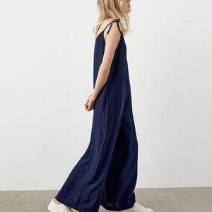 May include: Navy blue jumpsuit with wide legs and tie straps. The jumpsuit is made of a lightweight fabric and is paired with white sneakers. The model is walking in front of a white background.
