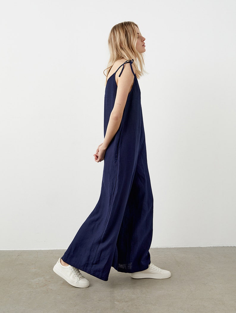May include: Navy blue jumpsuit with wide legs and tie straps. The jumpsuit is made of a lightweight fabric and is paired with white sneakers. The model is walking in front of a white background.