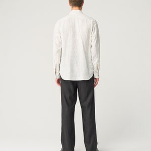 Op de afbeelding: Een persoon draagt een wit overhemd met verticale zwarte strepen en een donkergrijze broek. Het overhemd heeft lange mouwen en een kraag. De broek is wijd en lijkt van donkere denim te zijn. De persoon draagt zwarte schoenen.
