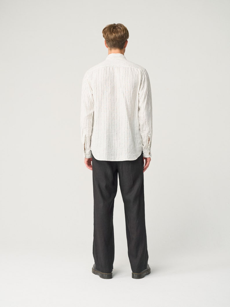 Op de afbeelding: Een persoon draagt een wit overhemd met verticale zwarte strepen en een donkergrijze broek. Het overhemd heeft lange mouwen en een kraag. De broek is wijd en lijkt van donkere denim te zijn. De persoon draagt zwarte schoenen.