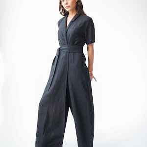 Könnte beinhalten: Dunkelgrauer Jumpsuit mit kurzen Ärmeln, einem Kragen und einem Wickelgürtel. Die weiten Hosenbeine sind bodenlang. Das Model trägt beige Spitzschuhe. Der Jumpsuit ist aus leichtem Stoff gefertigt.