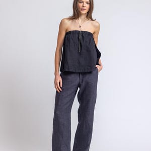 May include: A dark navy blue strapless top with a gold zipper and matching wide-leg pants. The outfit is made of a linen-like material. The model is wearing a gold necklace and black flats.