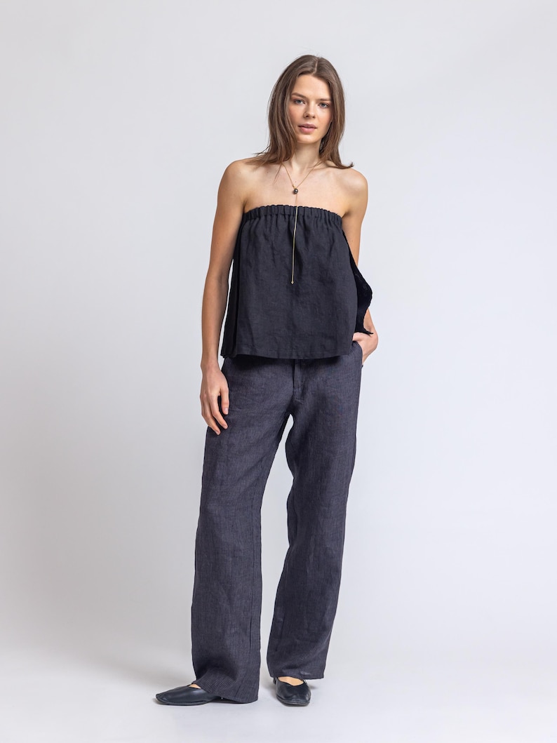 May include: A dark navy blue strapless top with a gold zipper and matching wide-leg pants. The outfit is made of a linen-like material. The model is wearing a gold necklace and black flats.