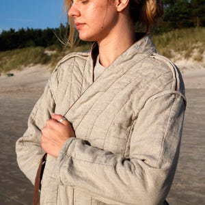 May include: A beige quilted jacket featuring vertical stitching, a high collar, and long sleeves. The garment has a dark brown inner lining and a matching brown tie belt visible at the side. The fabric has a textured, woven appearance.