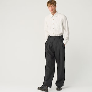 Op de afbeelding: Een persoon draagt een cr&egrave;mekleurig, gestreept button-down overhemd en een donkergrijze, geplooide broek. De outfit wordt gecompleteerd met zwarte leren schoenen. De kleding is minimalistisch gestyled.