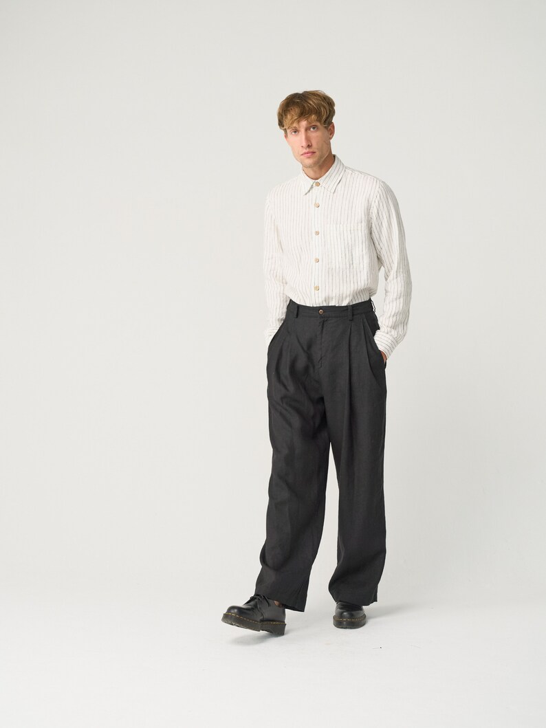 Op de afbeelding: Een persoon draagt een cr&egrave;mekleurig, gestreept button-down overhemd en een donkergrijze, geplooide broek. De outfit wordt gecompleteerd met zwarte leren schoenen. De kleding is minimalistisch gestyled.