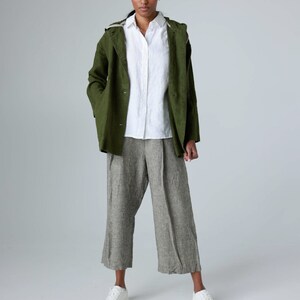 K&ouml;nnte beinhalten: Eine Frau tr&auml;gt eine gr&uuml;ne Jacke &uuml;ber einem wei&szlig;en Hemd mit Kn&ouml;pfen und einer grauen Leinenhose. Dazu wei&szlig;e Turnschuhe. Die Jacke hat eine Kapuze und die Hose ist weit geschnitten.