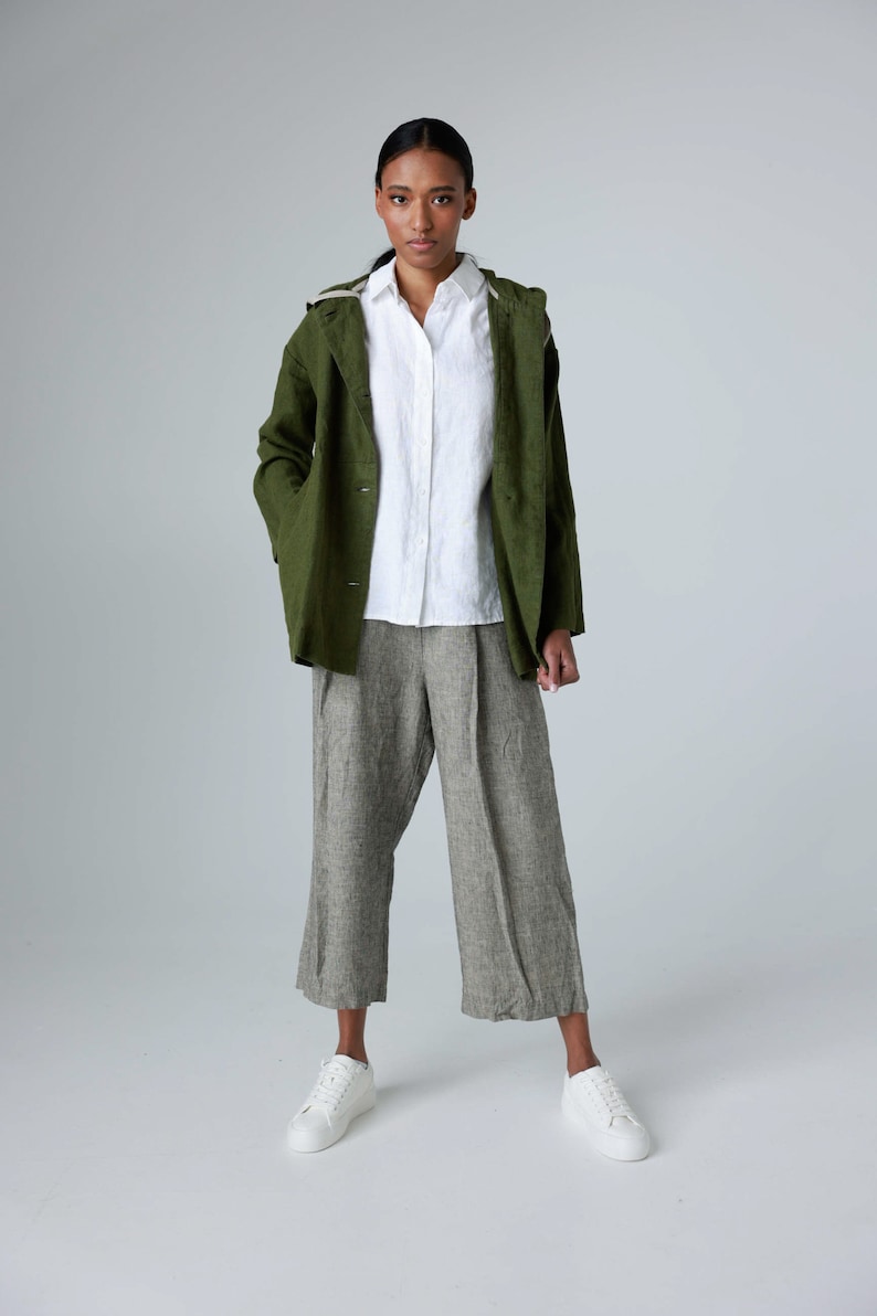 K&ouml;nnte beinhalten: Eine Frau tr&auml;gt eine gr&uuml;ne Jacke &uuml;ber einem wei&szlig;en Hemd mit Kn&ouml;pfen und einer grauen Leinenhose. Dazu wei&szlig;e Turnschuhe. Die Jacke hat eine Kapuze und die Hose ist weit geschnitten.