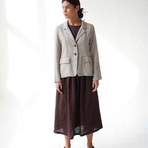 May include: A light beige linen blazer with two front pockets and brown buttons, paired with a dark brown midi skirt. The outfit is completed with beige flats. The ensemble is set against a white backdrop.