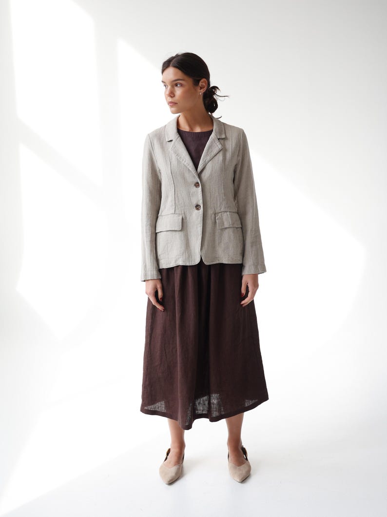 May include: A light beige linen blazer with two front pockets and brown buttons, paired with a dark brown midi skirt. The outfit is completed with beige flats. The ensemble is set against a white backdrop.