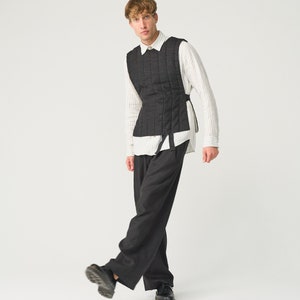 Op de afbeelding: Een zwarte gewatteerde vest met een verticaal stikselpatroon, gedragen over een gestreept overhemd en een donkergrijze broek. Het vest heeft een getailleerd silhouet en wordt vastgemaakt met zijbanden. De outfit wordt gecompleteerd met zwarte schoenen.