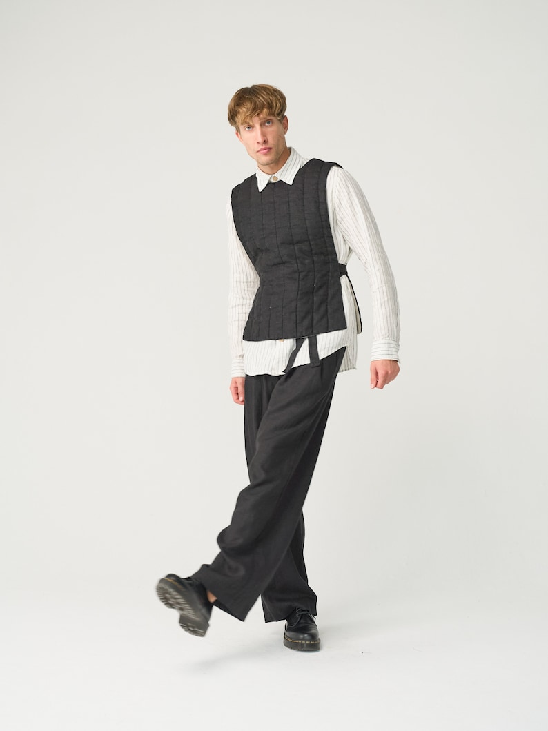 Op de afbeelding: Een zwarte gewatteerde vest met een verticaal stikselpatroon, gedragen over een gestreept overhemd en een donkergrijze broek. Het vest heeft een getailleerd silhouet en wordt vastgemaakt met zijbanden. De outfit wordt gecompleteerd met zwarte schoenen.