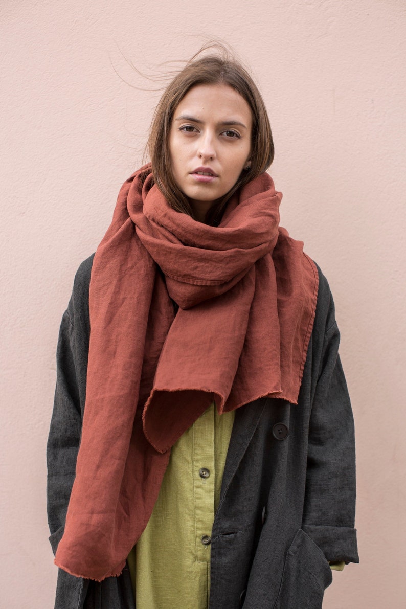May include: A rust-colored linen scarf is draped around the neck of a person wearing a dark gray coat and a light green shirt. The scarf is made of a soft, flowing fabric. The background is a solid, light pink wall.
