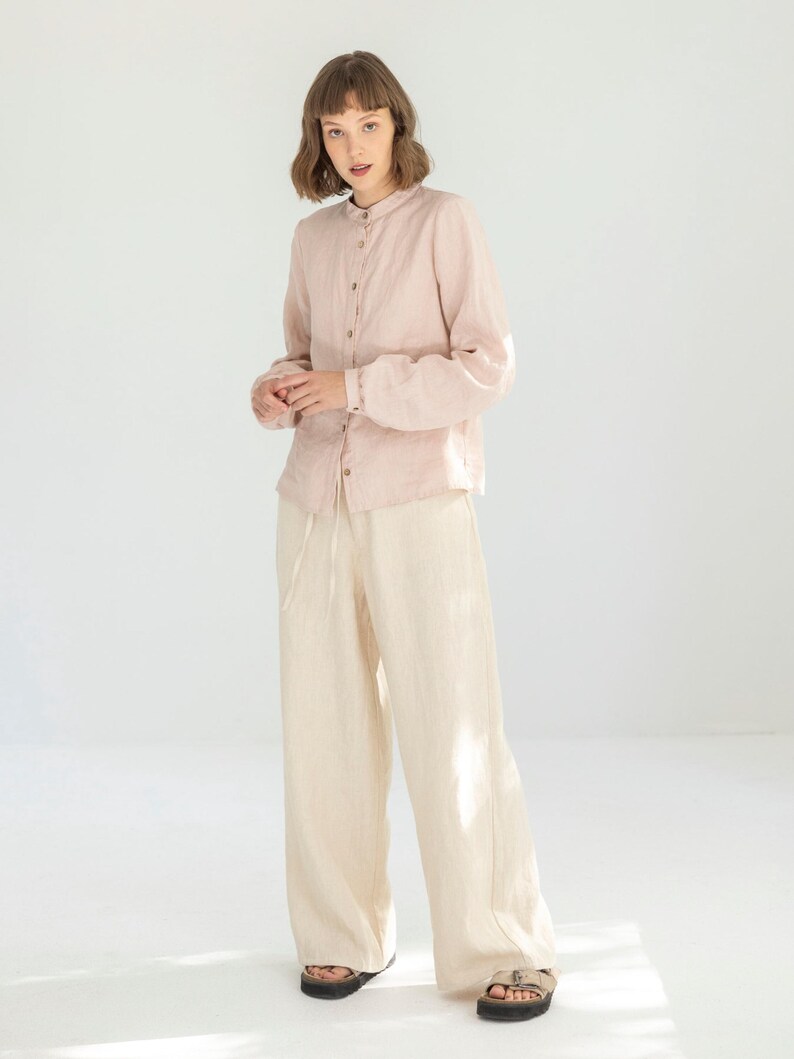 May include: A light pink linen shirt with a mandarin collar and button-down front is paired with cream-colored linen wide-leg pants. The outfit is completed with sandals. The clothing is made of a lightweight fabric.