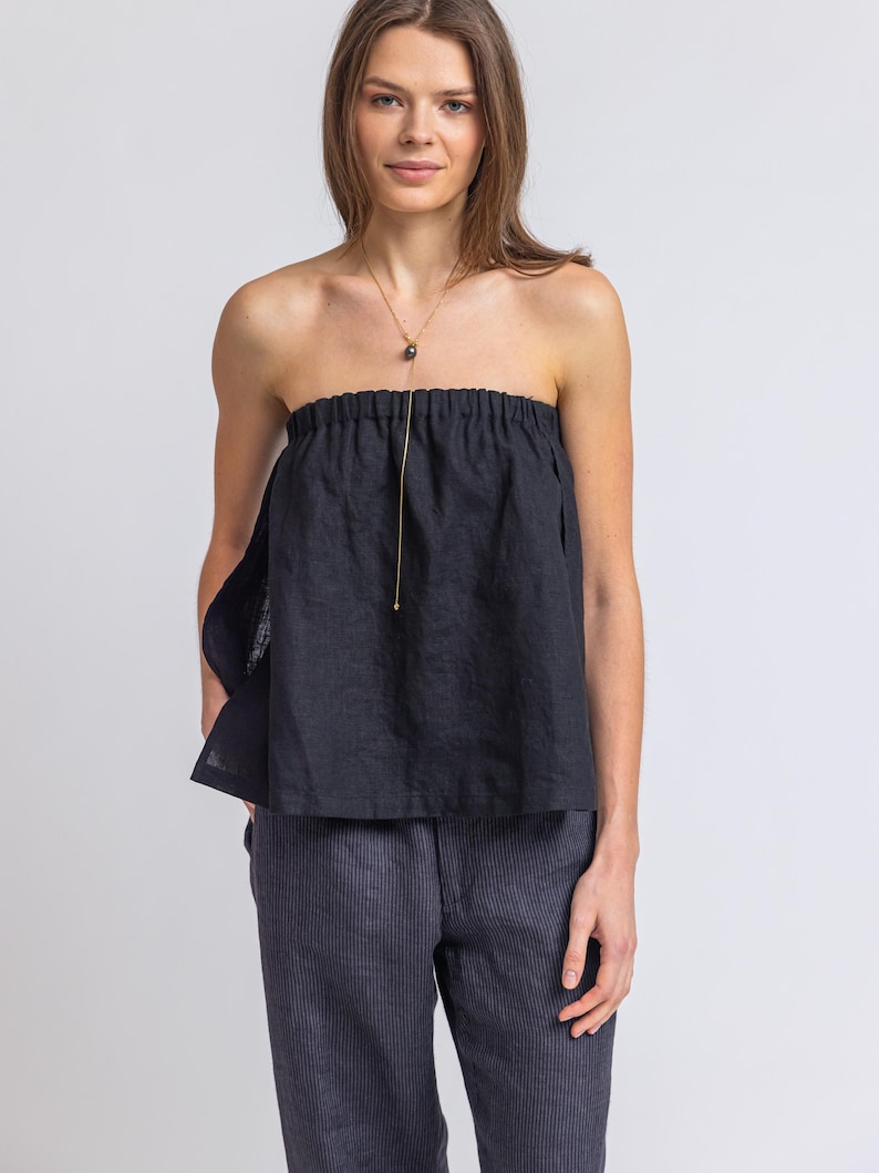 May include: A black strapless top with a gathered neckline and a gold necklace with a dark pendant. The top is paired with dark blue pinstripe pants. The outfit is simple and elegant.