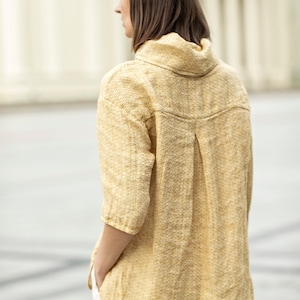 May include: A mustard yellow linen shirt with a high collar and three-quarter sleeves. The shirt has a textured weave and a relaxed fit. The model is wearing white pants. The shirt's design includes a pleat detail on the back.