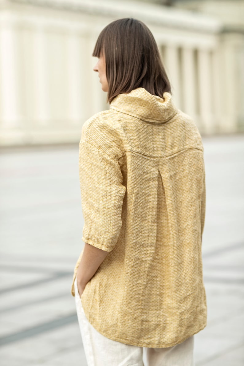 May include: A mustard yellow linen shirt with a high collar and three-quarter sleeves. The shirt has a textured weave and a relaxed fit. The model is wearing white pants. The shirt's design includes a pleat detail on the back.