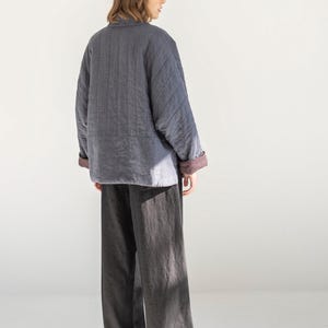 May include: A person wearing a grey quilted jacket with a burgundy lining and dark grey wide-leg trousers. The jacket has a quilted pattern and a collar. The trousers are a solid colour and have a loose fit. The outfit is simple and modern.
