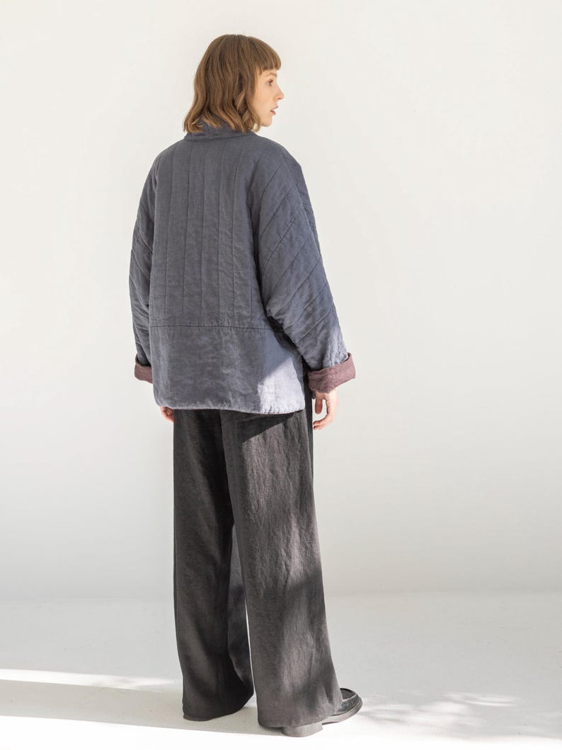 May include: A person wearing a grey quilted jacket with a burgundy lining and dark grey wide-leg trousers. The jacket has a quilted pattern and a collar. The trousers are a solid colour and have a loose fit. The outfit is simple and modern.