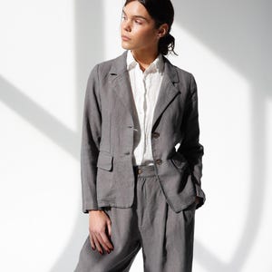 May include: A gray linen suit, consisting of a blazer and matching trousers, is worn over a white button-down shirt. The blazer has two buttons and a notched lapel. The trousers are high-waisted with a relaxed fit. The outfit is suitable for business or casual wear.
