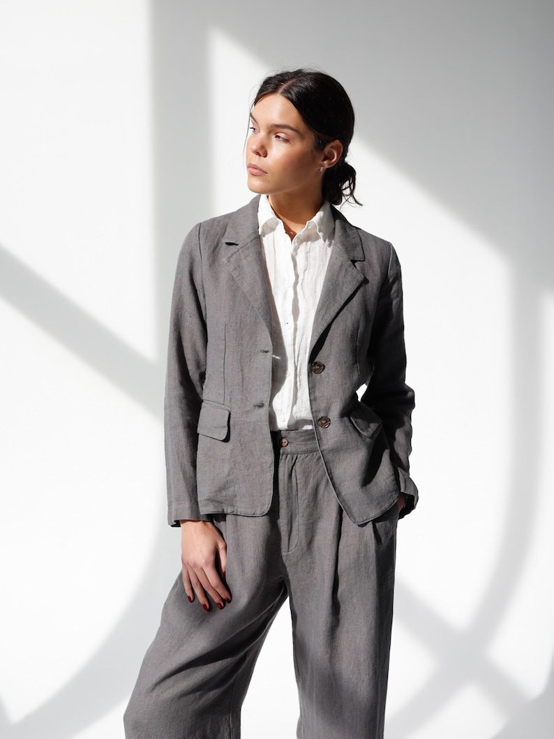 May include: A gray linen suit, consisting of a blazer and matching trousers, is worn over a white button-down shirt. The blazer has two buttons and a notched lapel. The trousers are high-waisted with a relaxed fit. The outfit is suitable for business or casual wear.